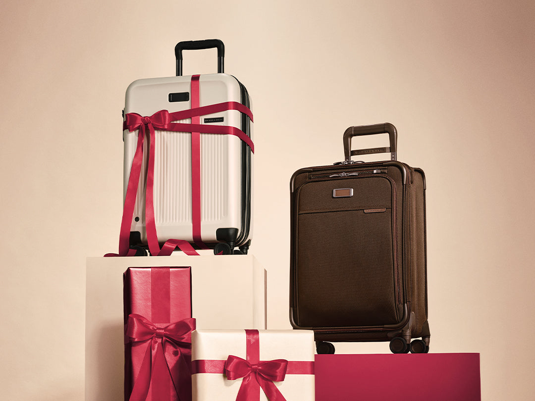 Two hard-shell suitcases, one silver with a bright pink ribbon and one dark brown, displayed next to wrapped gift boxes on a warm beige background.
