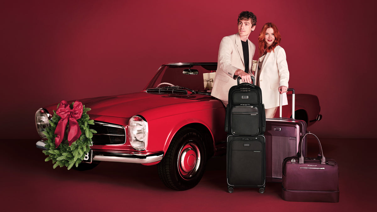 A man and a woman stand beside a red vintage convertible car decorated with a large red holiday bow and green wreath, with three hard-shell suitcases and a matching handbag arranged in front against a dark red studio backdrop.
