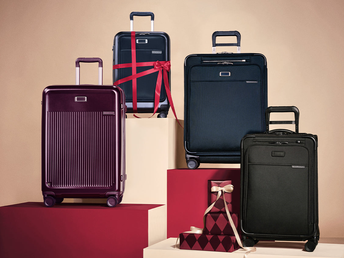 Four hard-shell and soft-sided carry-on suitcases in different colors are arranged on red and cream pedestals against a beige studio background. One suitcase is wrapped in a red ribbon, and two small gift boxes are placed in front.